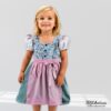 Ein Mädchen trägt das fertig genähte Modell aus dem Schnittmuster Kinderdirndl Resli im Garten. Zu sehen ist ein traditionelles Dirndl in Blau mit Blumenmuster und einer rosa Schürze.