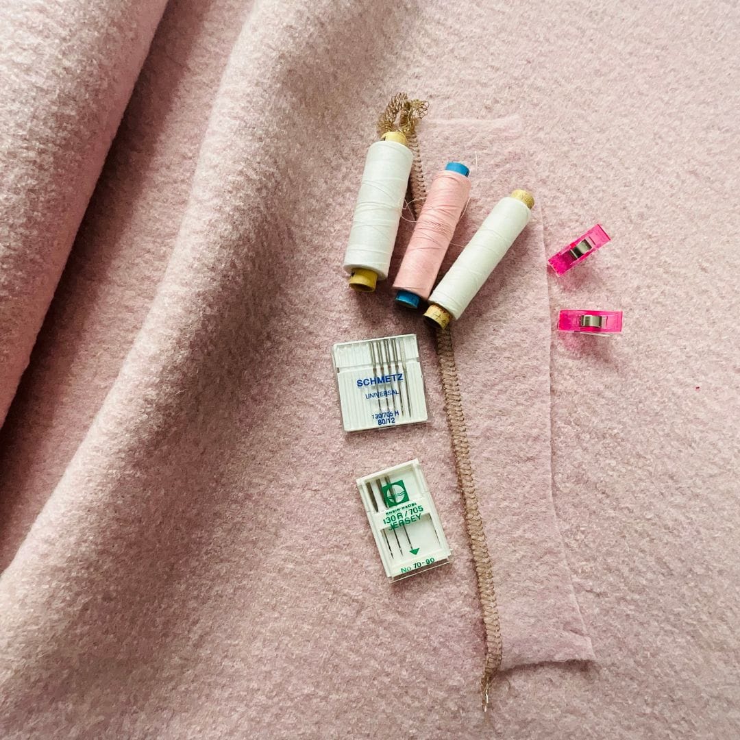 Zubehör zum Walkstoff nähen: Farblich passendes Garn, Nähmaschinennadeln und Stoffclips auf rosa Wollwalk.